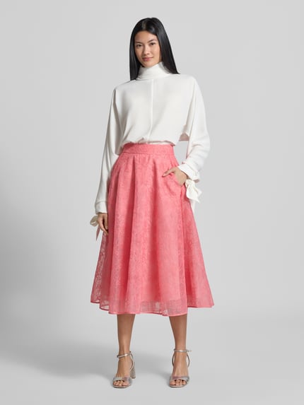Frauenmodel trägt Relaxed Fit Langarmbluse mit Rollkragen von Emporio Armani in Weiß, langärmliges Design mit Zierschleifen am Ärmelabschluss, kombiniert mit rosa weitem Rock, Ganzkörperansicht von vorne.