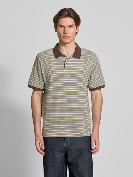 Męski model nosi koszulkę polo Regular Fit Poloshirt mit Label-Detail Modell 'Dion' od Carhartt Work In Progress z bawełny, wzór na całej powierzchni, detal z logo, listwa guzikowa, długość tyłu w rozmiarze M: 71 cm, krój regular fit, całkowity widok z przodu.