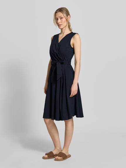 Kobiece model prezentuje sukienkę 'Kleid mit V-Ausschnitt in Wickel-Optik' od More & More, mieszanka bawełny, krój taliowany, dekolt w serek, kopertowy styl, bez rękawów, odpinana tasiemka w talii, długość tyłu w rozmiarze 36: 100 cm, szerokość w rozmiarze 36: 35 cm, ciemny granat, gładka tekstura, widok całej sylwetki z przodu.