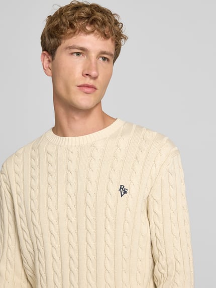 Męski model nosi Slim Fit Strickpullover reine Baumwolle od REVIEW, kremowa bawełna, ścieg warkoczowy, wyhaftowane logo, prążkowane ściągacze, zbliżenie na górną część ciała