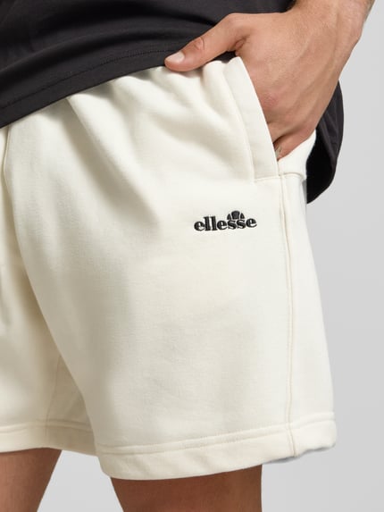 Mężczyznę model nosi Shorts mit Logo-Stitching Modell 'ZANICA' von Ellesse z bawełnianej mieszanki, kolor ecru, haftowane logo, kieszenie boczne, relaxed fit, szerokość w rozmiarze M 43 cm, długość nogawki od wewnątrz 18 cm, zbliżenie na górną część nóg