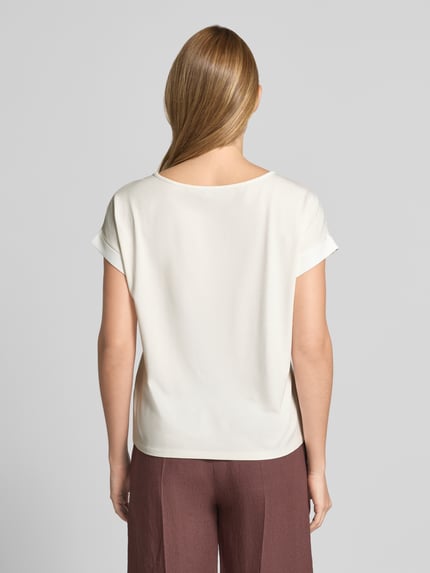 Vrouwenmodel draagt 'Blusenshirt mit verlängertem Rücken und Brusttasche' van monari in gebroken wit, recht model, ronde hals, opgerolde mouwen, borstzakje, verlengde achterkant, voorkant volledig glad, context: dames blusenshirt, ruglengte maat 36: 54 cm, mouwlengte: 3 cm, full body achteraanzicht.