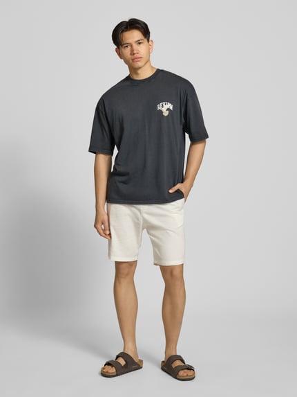 Männermodel trägt T-Shirt mit Logo- und Motiv-Print von REVIEW in Dunkelgrau, Reine Baumwolle, Loose-Fit, Gerippter Rundhalsausschnitt, Ganzkörperansicht von vorne.