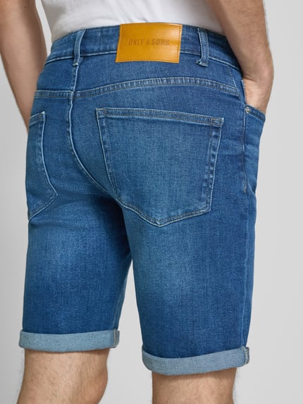 Männermodel trägt Regular Fit Jeansshorts aus Baumwoll-Mix Modell 'PLY' von Only & Sons, Denim-Optik, Label-Patch, Nieten, 5-Pocket-Design, Ganzkörperansicht Rückansicht.