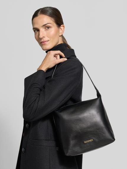 Vrouwenmodel draagt zwarte Handtasche met Label-Applikation von BOGNER met schouderband, binnenvak met rits, hoofvak met ritssluiting, breedte 31 cm, lengte 30 cm, diepte 15 cm, bandlengte 55 cm, labelapplicatie, ganzlichaanszicht van voren.