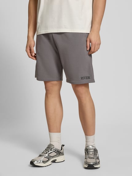 Männermodel trägt Regular Fit Shorts mit Label-Stitching von REVIEW aus mieszanka bawełny, krój regular fit, szary, elastyczny pas, wyhaftowane logo, szerokość w rozmiarze M: 44 cm, długość wewnętrzna nogawki w rozmiarze M: 22 cm, Ganzkörperansicht von vorne