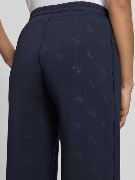 Männermodel trägt granatowe Wide Fit Sweatpants mit Allover-Logo-Print Modell 'Calista' von Guess Activewear, elastyczny materiał, widoczne logo, elastyczny pas z tunelowym ściągaczem, kontekst: Wide Fit, długość nogawki w rozmiarze S: 80 cm, detailaufnahme tylnej części spodni.