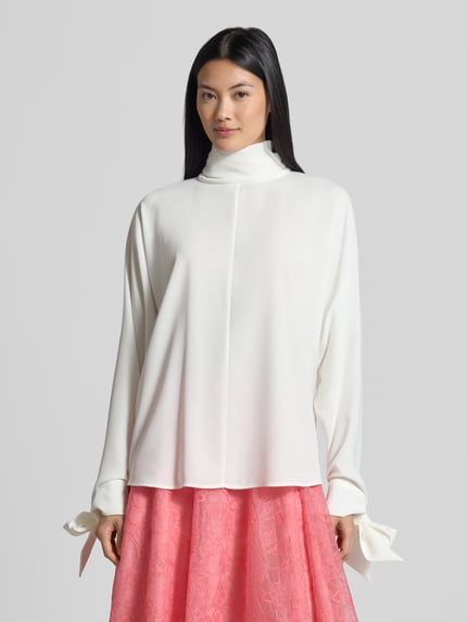 Frauenmodel trägt Relaxed Fit Langarmbluse mit Rollkragen von Emporio Armani in Weiß, leichte Stoffstruktur, Zierschleifen am Ärmel, Ganzkörperansicht von vorne.