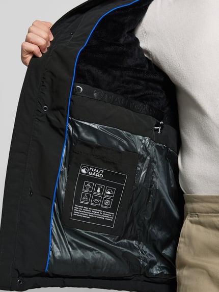 Männermodel trägt Parka mit Kapuze von NAUTGÅRD 'J-LHOTSE 226', schwarz, Label-Detail, Ziertasche, Reißverschlusstasche, Tunnelzug, Nahaufnahme Oberkörper