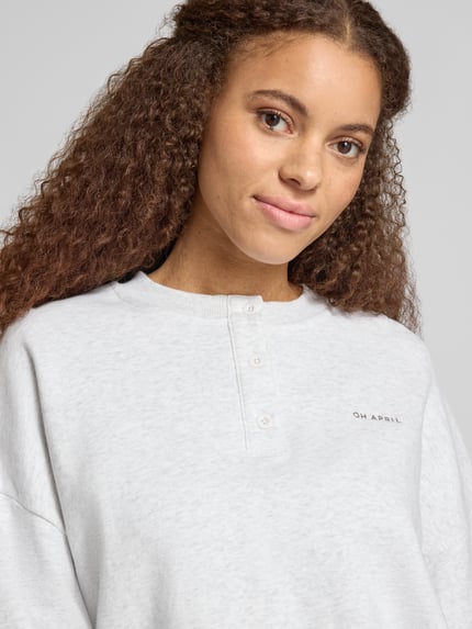 Frauenmodel trägt Boxy Fit Sweatshirt mit Knopfleiste Modell 'Aven' von OH APRIL aus reiner bawełna mit okrągły dekolt, listwa guzikowa, wyhaftowane logo, jasnoszary kolor, miękka faktura, Nahaufnahme Oberkörper.