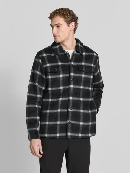 Männermodel trägt Hemdjacke mit Druckknopfleiste von BOSS Orange, kariertes Allover-Muster, Umlegekragen, Brusttasche, Ganzkörperansicht von vorne.
