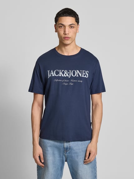 Męski model nosi t-shirt z bawełny model 'DEVIN' od Jack & Jones, prosty krój, okrągły dekolt, nadruk z logo, długość tyłu w rozmiarze M: 65 cm, kolor granatowy, gładka faktura, widok całej sylwetki z przodu.