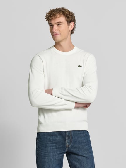Männermodel trägt weißen Regular Fit Pullover aus Baumwoll-Mix von Lacoste, kombiniert mit blauer Jeans, Ganzkörperansicht von vorne. Herren Pullover von Lacoste