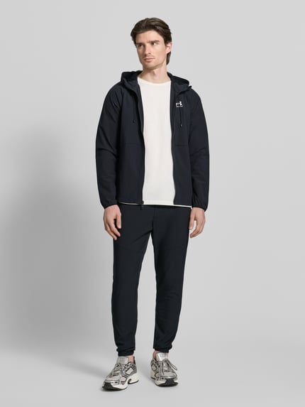 Männermodel trägt Jacke mit Kapuze von Under Armour in Schwarz mit Reißverschluss, Label-Print und lässigem Schnitt, Ganzkörperansicht von vorne.