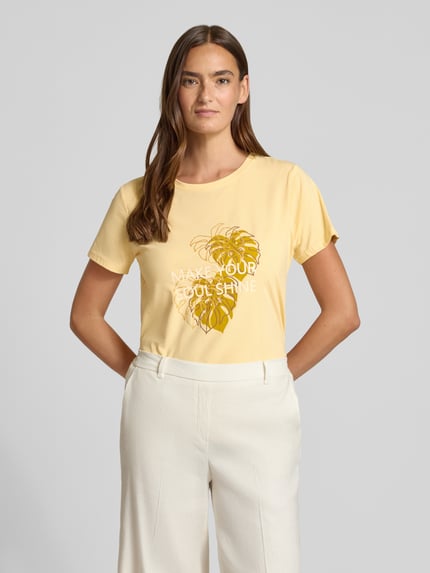 Vrouwenmodel draagt T-shirt met statement- en motiefprint model 'RINA' van Soyaconcept, puur katoen, regular fit, ronde hals, dames, ruglengte bij maat S 60 cm, geel met witte en gouden print, gladde textuur, voorkant, halve lichaamsopname.