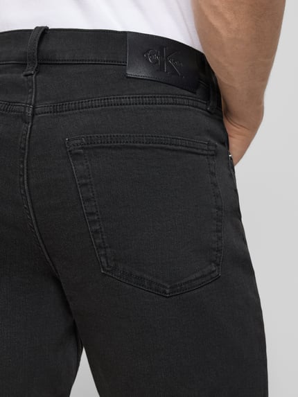 Mężczyzna nosi Slim Fit Jeansshorts aus Baumwoll-Mix Modell 'CK ESSENTIAL BLACK' von Calvin Klein Jeans, denim czarny z label patch, 5 kieszeni, guzik i ukryty zamek, krój slim fit, bawełniany materiał, detal z paskiem; zbliżenie na tylną kieszeń