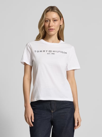 Frauenmodel trägt Regular Fit T-Shirt aus reiner Baumwolle von Tommy Hilfiger in Weiß, unifarben, Logo-Schriftzug, Rundhals, Kurzarm, Jersey-Textur, Nahaufnahme Oberkörper.
