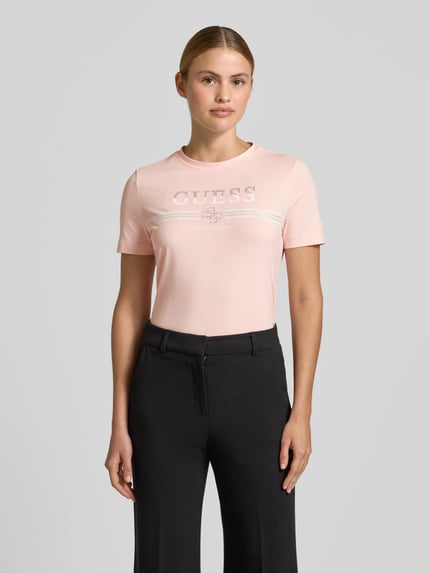 Frauenmodel trägt T-Shirt mit Strasssteinbesatz Modell 'SIENA' von Guess, rosa, Logo-Print, Baumwoll-Mix, Ganzkörperansicht von vorne.