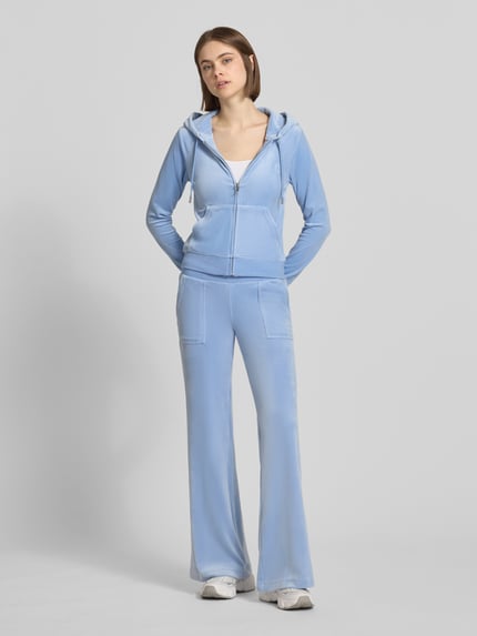 Frauenmodel trägt Low Rise Sweatpants mit Eingrifftaschen Modell 'LAYLA' von Juicy Couture, Damen Sweatpants aus Material-Mix mit Stretch-Anteil, Elastikbund, hellblau, glatt mit leichten Nähten, Ganzkörperansicht von vorne. Innenbeinlänge bei Größe S: 82 cm.