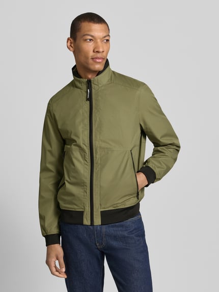 Männermodel trägt Regular Fit Jacke mit Stehkragen von MCNEAL in Olivgrün mit Reißverschluss und elastischen Abschlüssen, Ganzkörperansicht von vorne, Rückenlänge bei Größe M 64 cm, Schulterbreite 43 cm, Brustweite 55 cm, Ärmellänge 67 cm.