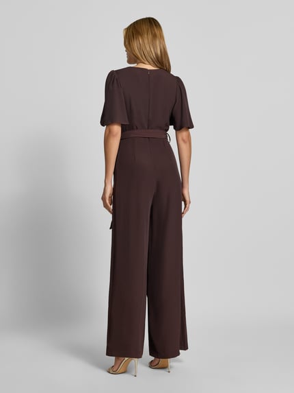 Frauenmodel trägt Jumpsuit mit Stoffgürtel von Rinascimento, braune Farbe, lockerer Material-Mix mit Stretch, tailliert, Kurzarm, Ganzkörperansicht Rückansicht. Damen Jumpsuit, V-Ausschnitt, Raffungen, Rückenlänge S 148 cm, Brustweite S 37 cm, Ärmellänge S 30 cm, Gesamtlänge S 147 cm, Innenbeinlänge S 79 cm.