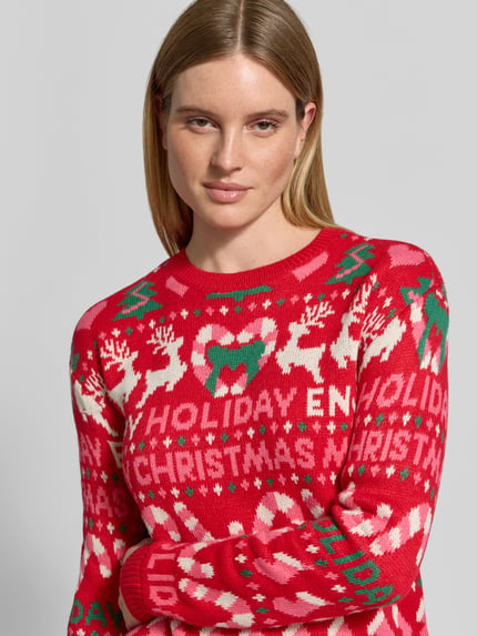 Frauenmodel trägt Pullover mit Weihnachtsmotiv-Frontprint von VERO MODA, rot, grün und weiß, Strickoptik, grafischer Allover-Muster, Nahaufnahme Oberkörper.