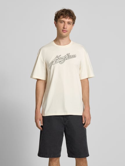 Męski model nosi Oversized T-Shirt mit Logo-Stitching von KARL KANI, czysta bawełna, krój oversized fit, okrągły dekolt, długość tyłu w rozmiarze M: 73 cm, jasnokremowy z czarnym logo, widok całej sylwetki od przodu.