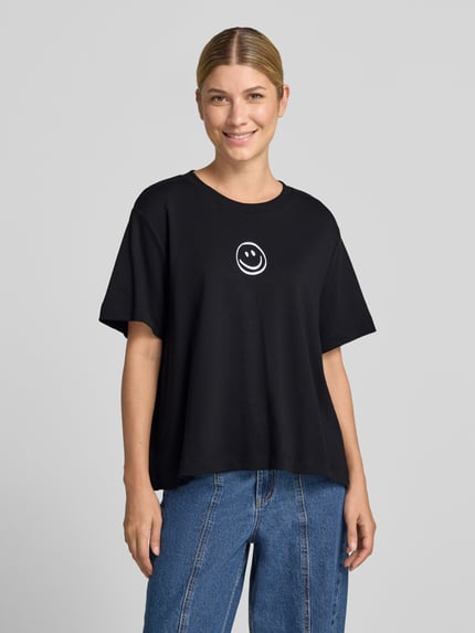 Frauenmodel trägt Boxy Fit T-Shirt aus reiner Baumwolle Modell 'SARA' von VERO MODA, schwarz mit weißem Smiley, Ganzkörperansicht von vorne.