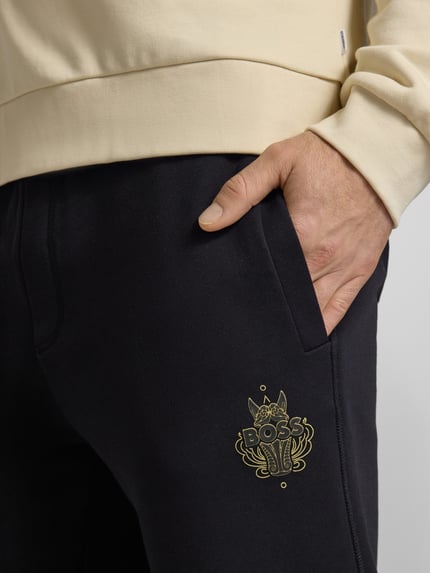 Männermodel trägt Oversized Jogginghose aus reiner Baumwolle Modell 'WEI_CNY' von BOSS in Schwarz mit goldfarbenem Label Print, Sweatstruktur, seitliche Eingrifftaschen; Nahaufnahme Oberkörper