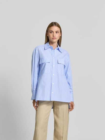 Frauenmodel trägt Relaxed Fit Hemdbluse aus Baumwolle von Joseph, hellblau-weiß gestreift, durchgehende Knopfleiste, Langärmelig, Ganzkörperansicht von vorne. Rückenlänge bei Größe 36: 83 cm, Ärmellänge: 58 cm, verlängerte Rückseite, glatte Struktur.