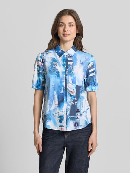 Modelka nosi Regular Fit Hemdbluse mit Allover-Motiv-Print von Marc Cain z bawełny, kołnierzyk koszulowy, krótki rękaw, elastyczne zakończenia rękawów, długość tyłu 62 cm w rozmiarze 36, długość rękawa 28 cm w rozmiarze 36, nadruk z motywem na całej powierzchni, widok całej sylwetki z przodu.