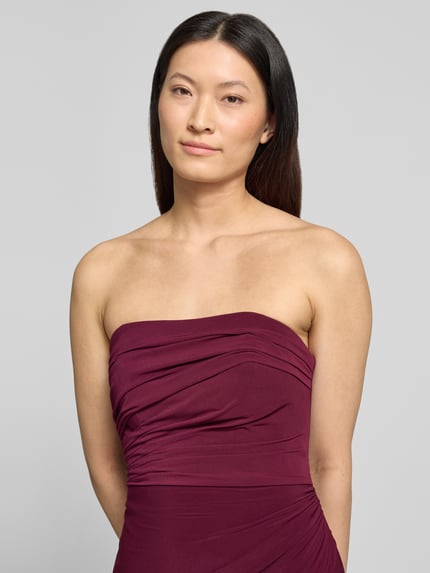 Frauenmodel trägt Slim Fit Abendkleid in Maxilänge mit Gehschlitz von Hey Kyla im Elastischer-Material-Mix, Schulterfrei, Raffung, Nahaufnahme Oberkörper. Rückenlänge bei Größe 36: 132 cm, Brustweite: 28 cm. Farbe: dunkles Bordeaux.
