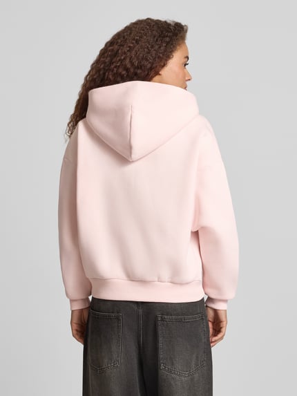 Vrouwenmodel draagt Relaxed Fit Sweatjacke mit Kapuze van REVIEW, lichtroze katoen met capuchon, logo-print, kangoeroezakken, relaxte fit, rugaanzicht.