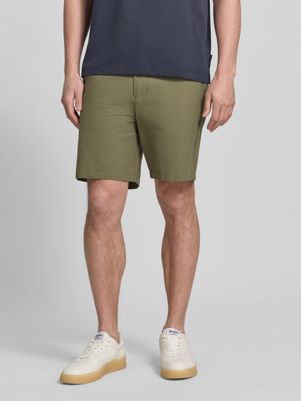 Männermodel trägt Regular Fit Bermudas aus Baumwoll-Mix Modell 'KARL' von SELECTED HOMME, wzór w paski, wykończenie materiału: seersucker, kolor oliwkowy, wpuszczane i tylne kieszenie, szlufki na pasek, zapięcie na guzik i zakryty zamek błyskawiczny, elastyczny ściągacz, krótka długość, zdjęcie: całkowity widok z przodu