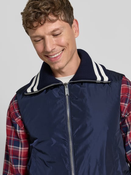 Männermodel trägt Weste mit Kontraststreifen von Dsquared2, marineblau mit weißem Kragen, Zweiwege-Reißverschluss, Nahaufnahme Oberkörper. Gerade Passform, Umlegekragen in Ripp-Optik, elastischer Bund, kombiniert mit kariertem Hemd.