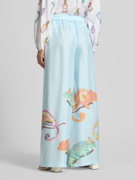 Vrouwenmodel draagt Wide Fit Seidenhose met Motief-Print van HERZENS in lichtblauw met zachte kleuren, zuiver zijde, steekzakken, elastische band met tunnelkoord, motiefprint, bandwijdte maat 36: 36 cm, binnenbeenlengte maat 36: 78 cm, wide fit, ganzebenenaanzicht achteraan.