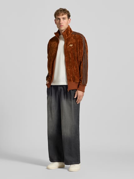Männermodel trägt Sweatjacke mit Samtoptik Modell 'FIREBIRD TT VEL' von adidas Originals, braun, glänzend, Logo, Kontraststreifen, Ganzkörperansicht von vorne.