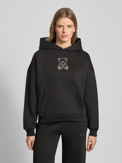 Frauenmodel trägt schwarzen Hoodie mit Kapuze von Review, Ziersteinbesatz, Känguru-Tasche, Baumwoll-Mix, lockerer Schnitt, Ganzkörperansicht von vorne.