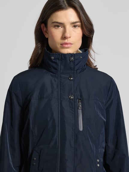 Modelka kobieca nosi Funktionsjacke mit Brusttasche od Gil Bret w kolorze granatowym, z wysokim kołnierzem, kapturem, taliowany krój, kieszonka na piersi z suwakiem, długość tyłu w rozmiarze 36: 74 cm, szerokość w ramionach w rozmiarze 36: 41 cm, szerokość w biuście w rozmiarze 36: 47 cm, długość rękawa w rozmiarze 36: 65 cm, prosta faktura tkaniny, zbliżenie na górną część ciała