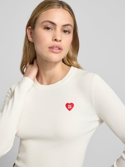 Modelka kobieca nosi Slim Fit Strickshirt aus Viskose-Mix Modell 'SHARPA' od HUGO, biały, długi rękaw, ribb, crew neck, naszywka, zbliżenie na górną część ciała.