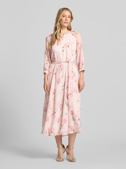 Vrouwenmodel draagt midi-jurk met strikceintuur van More & More, roze bloemenprint, getailleerd model, ronde hals met cut-out, 3/4-mouw, druppelvormige hals en ritssluiting achter, ruglengte maat 36: 128 cm, borstwijdte 42 cm, mouwlengte 58 cm, geheel van voren.
