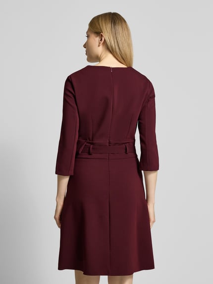 Frauenmodel trägt knielanges Cocktailkleid mit V-Ausschnitt von Rinascimento, aus Material-Mix mit Viskose-Anteil, taillierter Schnitt, Taillengürtel, 3/4-Ärmel, Nahtreißverschluss, Rückenlänge bei Größe S 92 cm, Brustweite 44 cm, Ärmellänge 45 cm; einfarbiges Bordeauxrot, strukturierte Oberfläche, Ganzkörperansicht Rückansicht.
