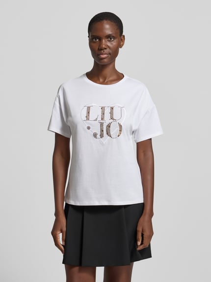 Kobiece model prezentuje Regular Fit T-Shirt mit Strasssteinbesatz von Liu Jo White z bawełny, logo, okrągły dekolt, długość tyłu 60 cm, Ganzkörperansicht von vorne.