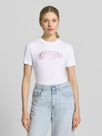 Frauenmodel trägt T-Shirt aus Viskose-Elasthan-Mix mit Logo von Guess, weiß mit rosa Schrift, Regular Fit, Rundhalsausschnitt, Nahaufnahme Oberkörper.