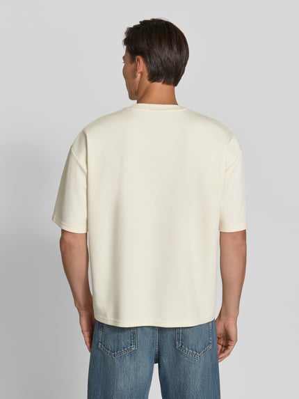 Männermodel trägt Oversized T-Shirt mit überschnittenen Schultern von REVIEW, cremeweiß, glatte Textur, lockerer Schnitt, Ganzkörperansicht Rückansicht. Rückenlänge bei Größe M: 70 cm, elastische Abschlüsse, Rundhalsausschnitt.
