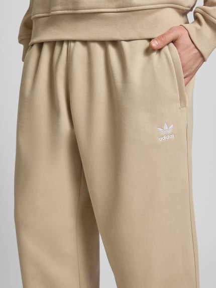 Männermodel trägt Wide Fit Sweatpants mit elastischem Bund von adidas Originals in Beige mit Logo-Stitching, Nahaufnahme Hüfte bis Oberschenkel. Baumwoll-Mix, sportlicher Look.