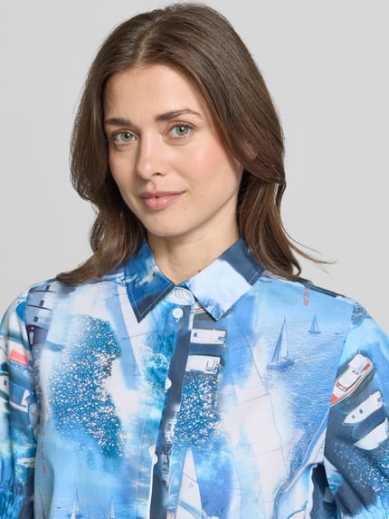 Kobiece modelka nosi Regular Fit Hemdbluse mit Allover-Motiv-Print von Marc Cain z bawełny, kołnierzyk koszulowy, z krótkim rękawem, elastyczne zakończenia rękawów, nadruk z motywem na całej powierzchni, niebieskie tonacje, gładka faktura, długość tyłu 62 cm, długość rękawa 28 cm, ujęcie: zbliżenie na górną część ciała.