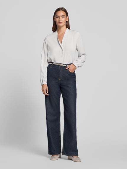 Kobiece modelka nosi Regular Fit Bluse mit V-Ausschnitt von Marc Cain, krój regular fit, dekolt w serek, detal z logo, długość tyłu w rozmiarze 36: 65 cm, długość rękawów w rozmiarze 36: 59 cm, jasna bluzka, ciemne spodnie, Ganzkörperansicht von vorne.