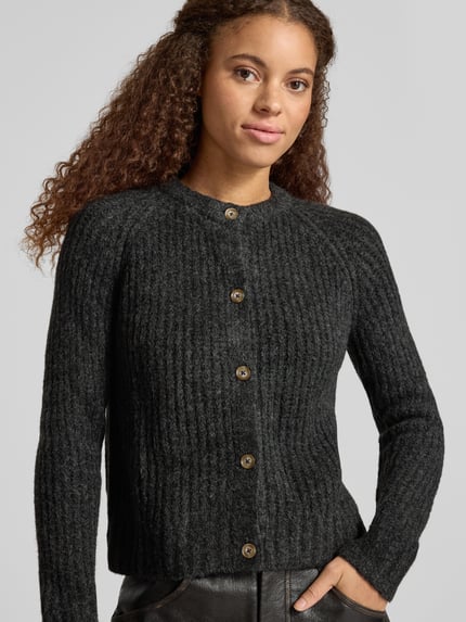 Modelka kobieca ma na sobie jednokolorowy, czarny kardigan rozpinany Regular Fit Strickjacke mit Woll-Anteil Modell 'ASHLEY CARDIGAN' od Pepe Jeans o fakturze dzianinowej ze strukturą, ozdobne guziki, długi rękaw raglanowy, skrócony krój, okrągły dekolt, prążkowane zakończenia; drobny splot, przód zapinany na guziki, materiał z dodatkiem wełny, widoczne przeszycia, widok całej sylwetki z przodu.