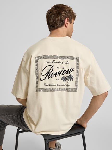 Mężczyznamodel nosi T-Shirt mit Rundhalsausschnitt und Label-Print od REVIEW, męski t-shirt z bawełny, krój relaxed fit, okrągły dekolt, nadruk z logo, długość tyłu w rozmiarze M: 71 cm, jasny kolor, prostokątny nadruk z logo na plecach, tekstura gładka, całkowity widok od tyłu.